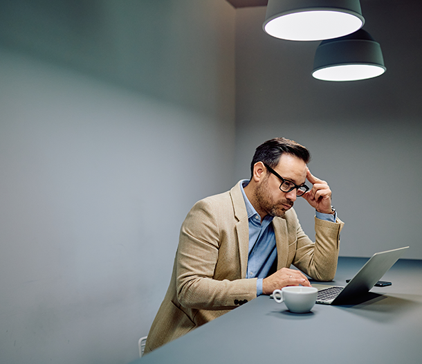 Man in a blazer looking stressed while working on a laptop at a table with a coffee cup, reflecting challenges in IT compliance and data security for small businesses.