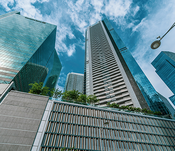 Cityscape with tall glass skyscrapers against a blue sky, viewed from street level with a decorative fence and greenery in the foreground