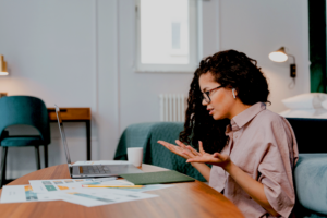 Woman with curly hair and glasses sits at a table with a laptop, gesturing with her hands during work in a bright office.
