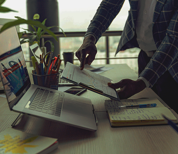 Person in a plaid jacket arranging papers at a desk with a laptop, tablet, notebook, and pens.
