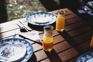 Outdoor wooden picnic table set with floral-patterned plates and two glass bottles of orange juice in sunlight.